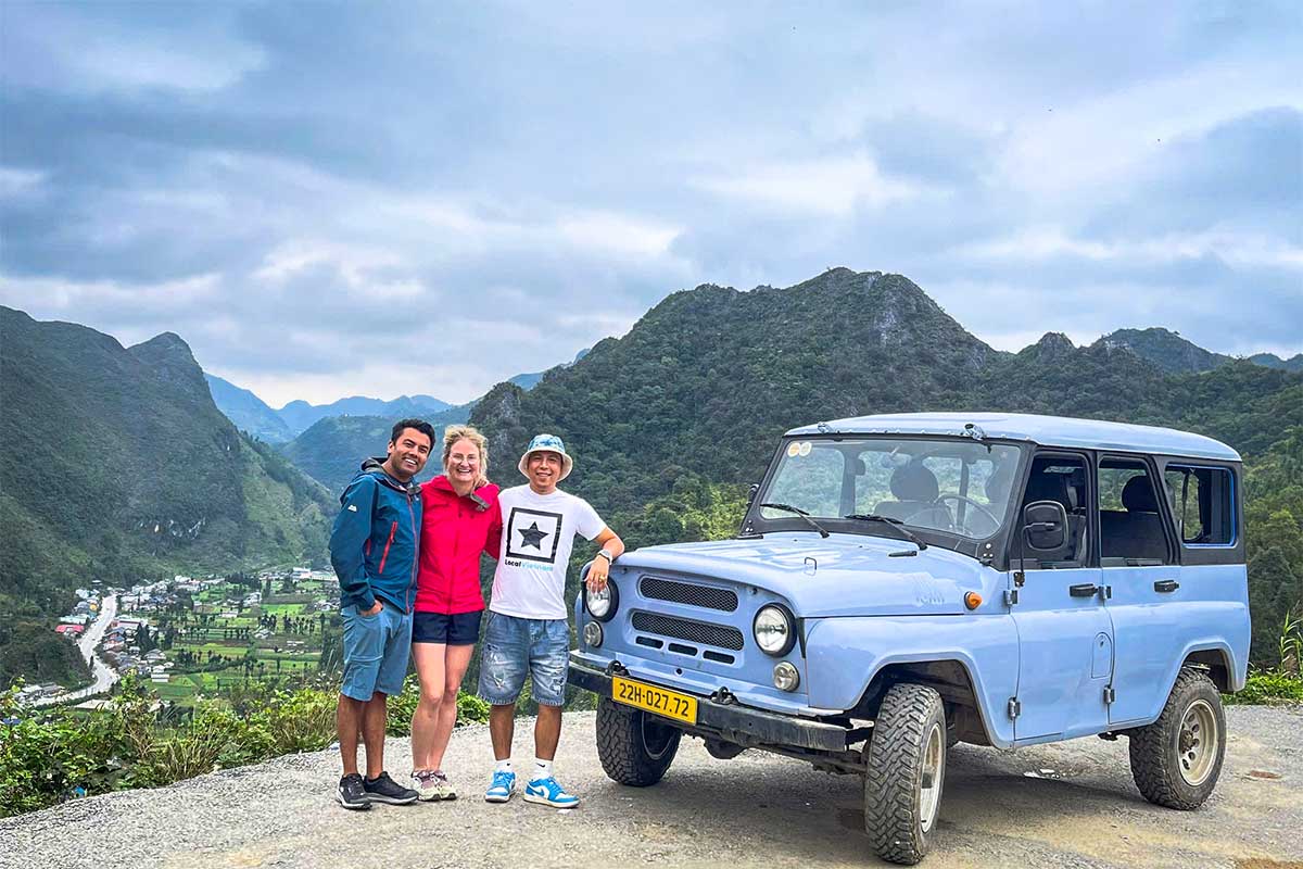 Drie reizigers staan lachend naast een lichtblauwe UAZ-jeep op een uitzichtpunt in Ha Giang, met valleien en karstbergen op de achtergrond.