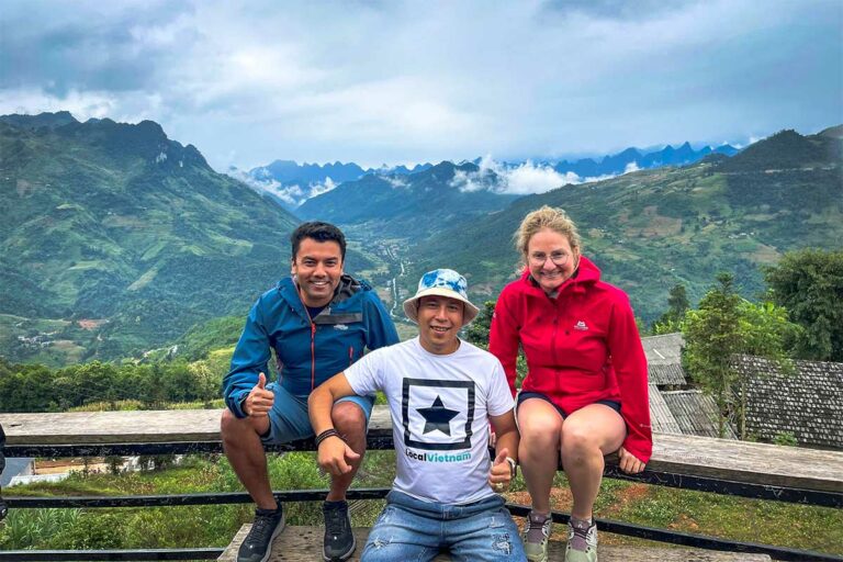 Een Local Vietnam-gids en twee reizigers zitten samen op een houten bankje met panoramisch zicht op een groene vallei en bergtoppen in Ha Giang.