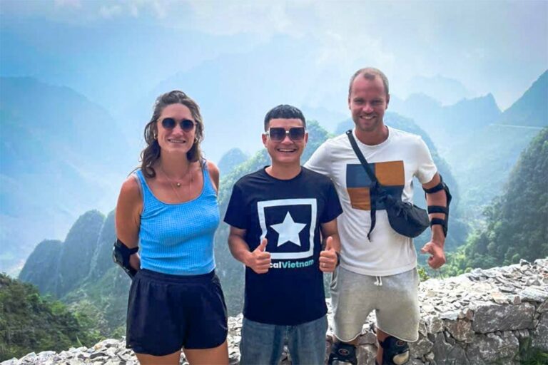 Twee reizigsters poseren met hun Local Vietnam-gids tijdens de Ha Giang Loop Easy Rider tour, met motorbikes en een helder berglandschap op de achtergrond.