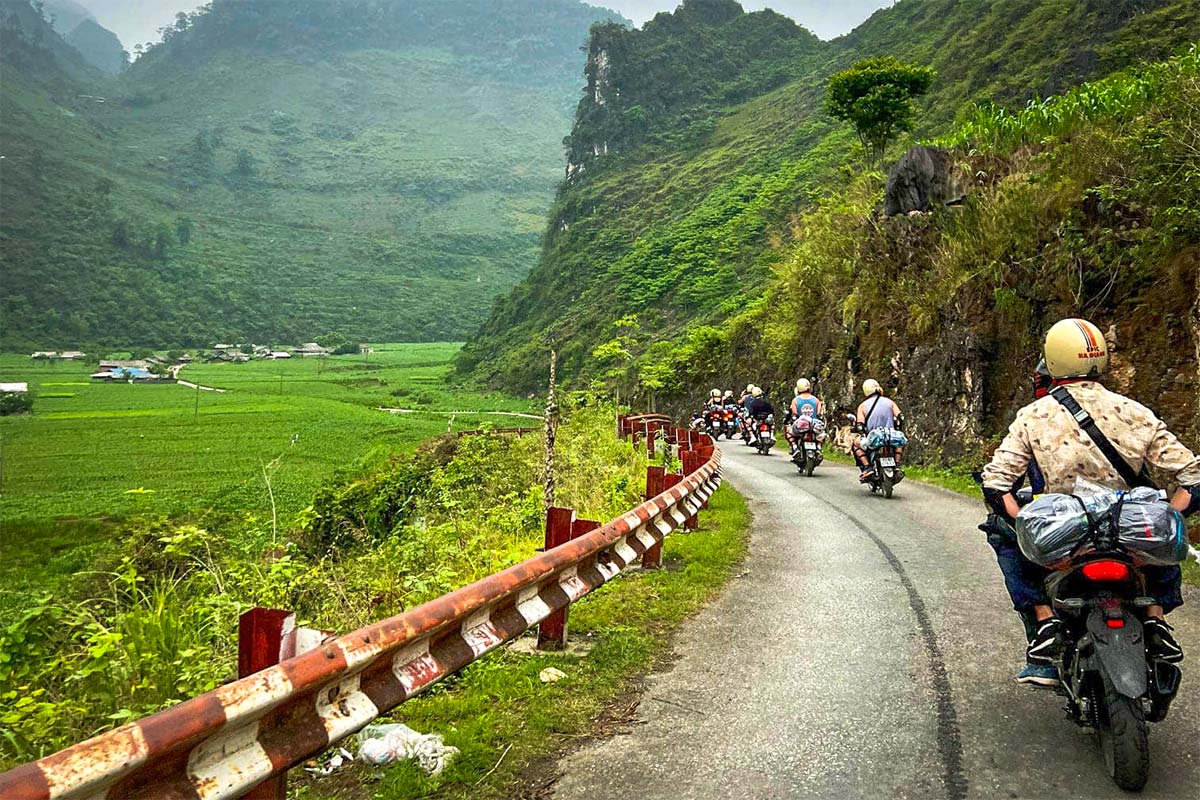 Een groep reizigers rijdt met hun Easy Rider-gidsen door een groene vallei tijdens de Ha Giang Loop groepstour, over een smalle weg langs rotswanden en rijstvelden.