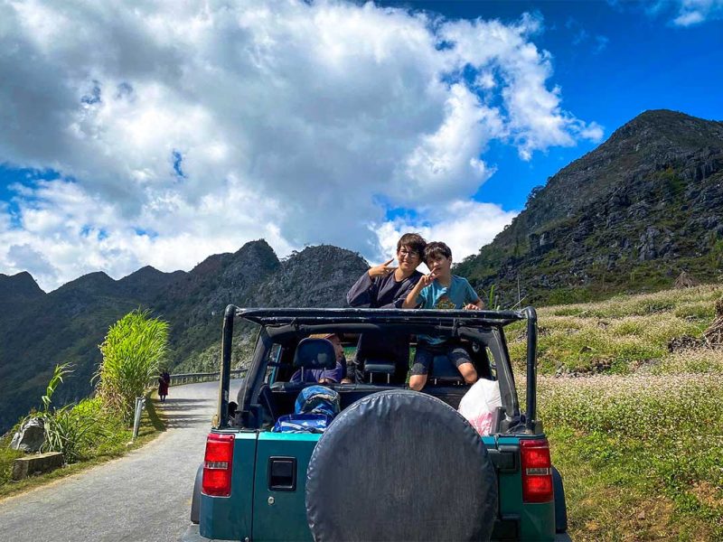 Jeep tour op de Ha Giang Loop met twee reizigers bovenop de open jeep, met uitzicht op steile kalksteenbergen en blauwe lucht.