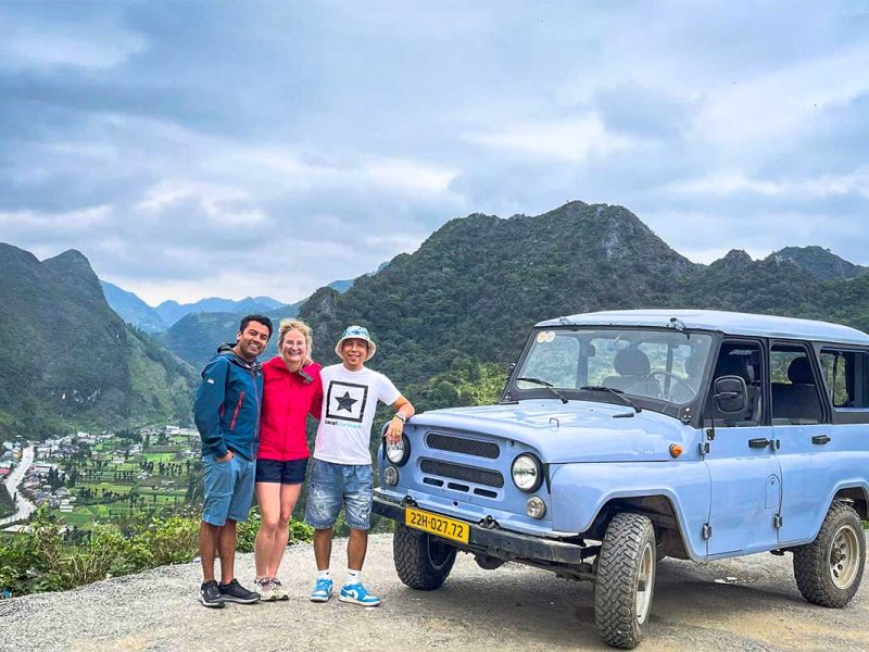Drie reizigers staan lachend naast een lichtblauwe UAZ-jeep op een uitzichtpunt in Ha Giang, met valleien en karstbergen op de achtergrond.