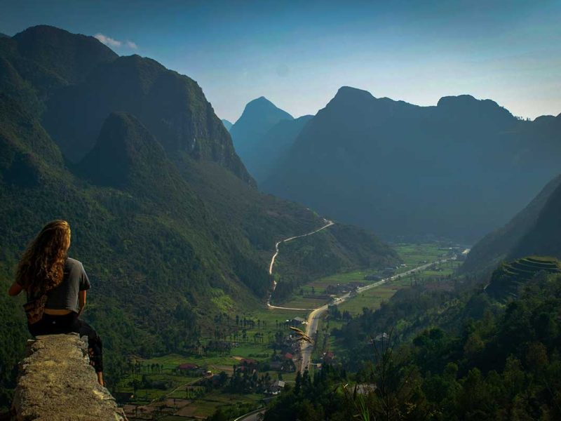 Reiziger die uitkijkt over de vallei en kronkelende bergwegen van de Ha Giang Loop bij zonsopkomst.